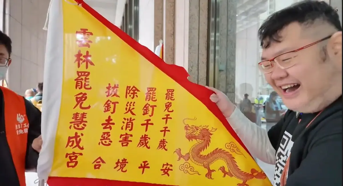 ▲四叉貓拿著雲林罷免慧成宮的令旗大笑，稱讚「好有中部人的fu」。（圖／翻攝四叉貓YT，2025.03.07）