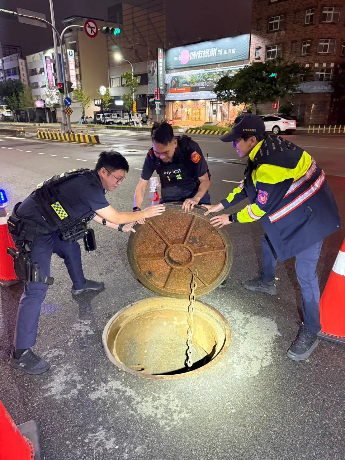 ▲三名警力頂著風雨使出吃奶力氣，才終於將「脫軌」的人孔蓋成功搬回原處，恢復交通通行（圖/記者鄧力軍翻攝）