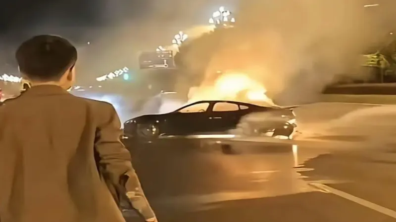 A black car is engulfed in flames with thick smoke rising during nighttime, while a person in a brown jacket watches nearby on a road that appears to be on Chengdu Tianfu Avenue.